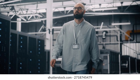 IT Engineer Walking Through Server Racks in Modern Data Center. Focused Man With Badge and Laptop Walking Among Rows of Glowing Server Racks. Concept of IT Infrastructure and Cloud Technology.