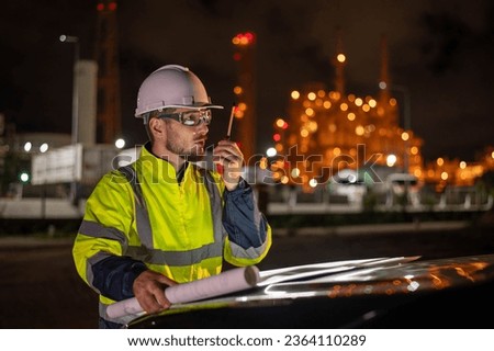 Engineer wear uniform and helmet stand in front of the car hand holding blueprint paper, survey inspection work plant site use radio communication to work with night lights oil refinery background.