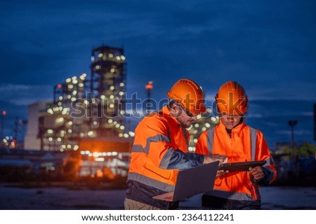 Engineer wear uniform and helmet stand workplace hand holding tablet and laptop computer, survey inspection team work plant site to work with night lights oil refinery background.