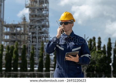 Engineer wearing safety helmet using walkie-talkie and tablet in industrial plant area, representing communication, safety management, inspection process and refinery oil plant factory operation.