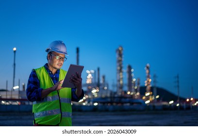 Engineer wearing safety uniform and helmet looking detail tablet on hand with oil refinery factory at night time background.