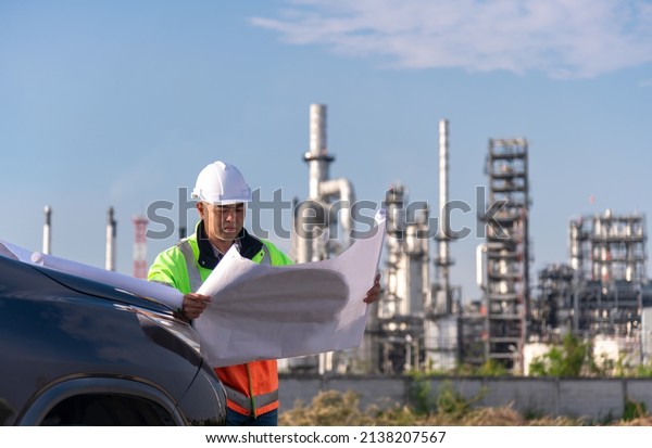 Engineer wearing uniform and helmet stand\
next to the car hand holding detail blue print paper, inspection\
and surveying work site progress with oil refinery power industrial\
factory  background.