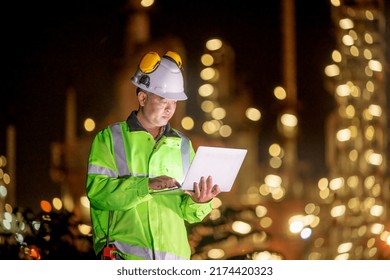 Engineer Wearing Uniform, White Helmet And Ear Muff Standing Hand Holding Detail Laptop Inspection Survey Work Plant Site Progress With Lighting Oil Refinery Industrial At Night Time  Background.