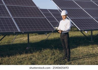 An engineer in a white hard hat inspects solar panels at a solar farm, demonstrating renewable energy efficiency and modern technology in a green, outdoor environment. - Powered by Shutterstock