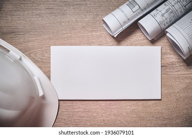 Engineer White Hardhat And Rolled Drawings With Copy Space. Blank White Envelope At Wooden Table Top Down View. Engineer Desk Workplace Mockup