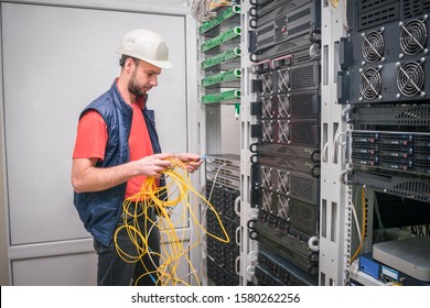 Engineer In A White Helmet Connects Internet Wires To New Equipment. Technology Concept. A Man Works In The Server Room Of A Data Center. A Technician Connects A Cable To A Stack Of Managed Switches