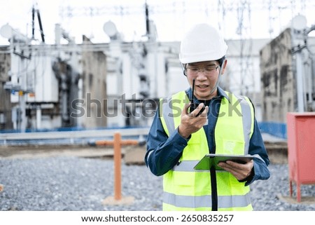 An engineer is working at high voltage power substation conducting inspection and check of infrastructure using walkie talkie and tablet for safety and communication