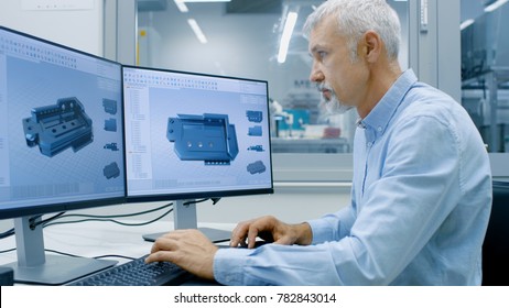 Engineer Working on a Personal Computer with Two Displays, He's Designing New Essential Component in CAD Program. Out of the Office Window Components Manufacturing Factory is Visible.