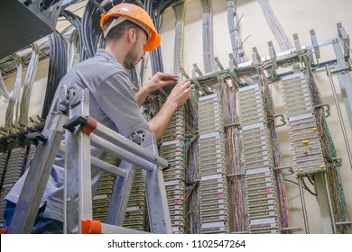  The Engineer Works In The Server Room Of The Data Center. Cable Laying In Telephone Exchange. A Worker Standing On The Stairs Commutes Telephone Wires.