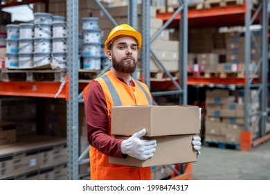 Engineer In Workwear Holding Boxes Against Racks With Goods