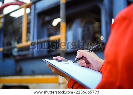 An engineer is writing on paper to list the maintenance detail with a heavy pumping machine as blurred background. Industrial expertise occupation working scene, selective focus at hand's part.