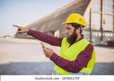 An Engineer With A Yellow Helmet On His Head And A Green Protective Vest Uses A Mobile Phone And Points His Finger. In The Background Is A Large Silo