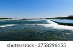 Engineered structure alters current just upstream of main falls. Birds gather along crest where water spills evenly. Background features forested shoreline and distant bridge spanning river valley.
