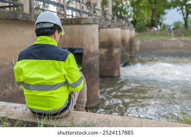 A engineering doing his checking routine. He is wearing hard hat and engineer uniform.Standing by the rail by the dam.Monitor water levels from the heavy rain that has been falling for several days.
