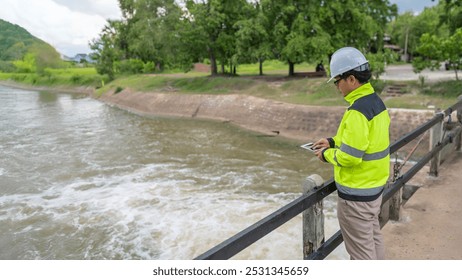 A engineering doing his checking routine. He is wearing hard hat and engineer uniform.Standing by the rail by the dam.Monitor water levels from the heavy rain that has been falling for several days.