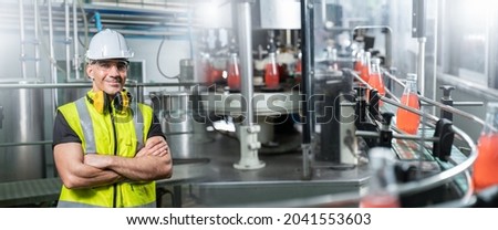Engineering male worker arms crossed wearing safety uniform and helmet in beverage factory industry,check quality of electric machine to produce bottle before distribution to market business.