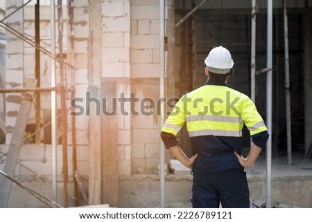 Engineering man turning back to camera looking at incomplete house constructed with two hand at waist, blured constuction brick and yellow light.
