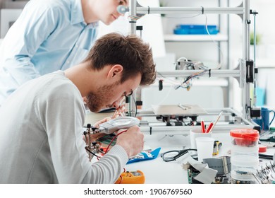 Ingenieurstudenten, die im Labor arbeiten, ein Student verwendet einen Spannungs- und Stromtester, ein anderer Student im Hintergrund verwendet einen 3D-Drucker