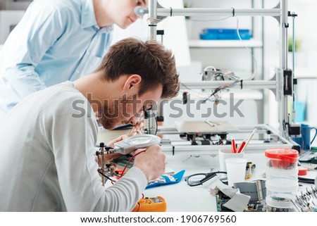 Engineering students working in the lab, a student is using a voltage and current tester, another student in the background is using a 3D printer