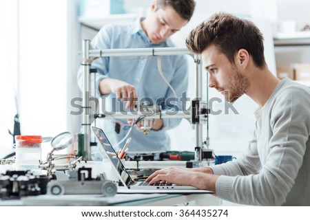 Engineering students working in the lab, a student is adjusting a 3D printer's components, the other one on foreground is using a laptop