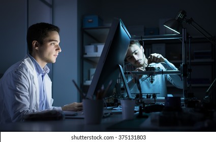 Engineering Team Working In The Lab At Night, A Student Is Working With A Computer And The Other One Is Adjusting A 3D Printer's Components