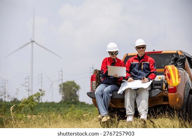 Engineers checking blueprint on pickup truck at wind turbine farm, working on renewable energy project planning and site inspection. Clean power, field operation, teamwork, sustainability concept.