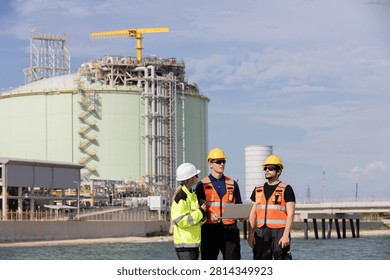 Engineers collaborating on construction site near industrial plant with crane and tanks in background