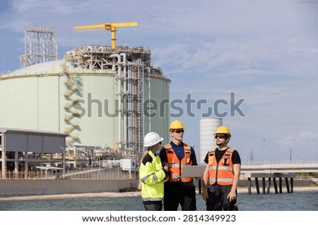Engineers collaborating on construction site near industrial plant with crane and tanks in background