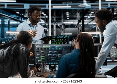 Engineers doing brainstorming, programming in server farm housing infrastructure storing datasets. Teamworking colleagues run code on PC to repair data center racks doing computational operations