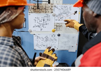 Engineers examining mechanical blueprints and discussing technical details, with tools and schematics pinned on wall for reference - Powered by Shutterstock