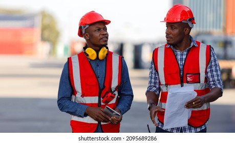 Engineers Specializing In Container Yard Management Are Working On Outdoors. Two African-American Container Yard Workers Holding Blueprint And Discuss The Placement Of In The Container Yard.