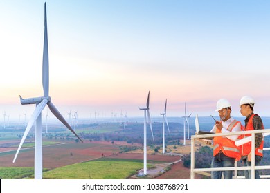 Engineers And Technician Looking At Computer For Repair And Control Working Assisted By Crane And Elevator At Wind Turbine Power Generator Station In Sunset
