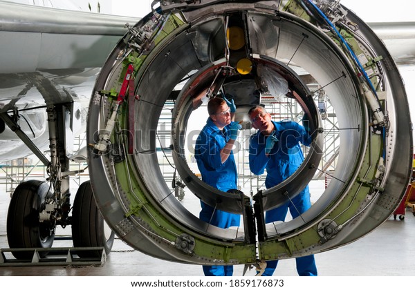 Engineers in uniforms inspecting the engine\
casing of a passenger jet at a\
hangar.