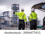 Engineers wear uniform standing near the open trunk of a vehicle hand holding blueprint paper show, survey inspection work plant site use tablet see detail of work with oil refinery background.