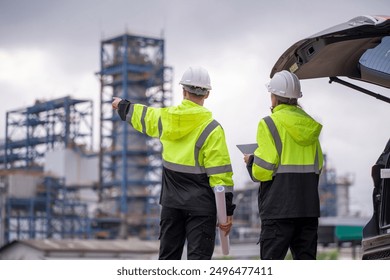 Engineers wear uniform standing near the open trunk of a vehicle hand holding blueprint paper show, survey inspection work plant site use tablet see detail of work with oil refinery background. - Powered by Shutterstock