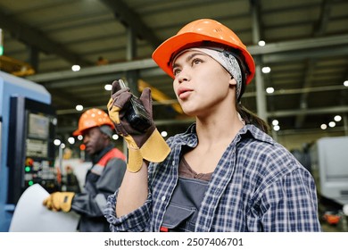 Engineers wearing safety helmets and gloves working on machinery in spacious industrial setting with focus on female engineer using radio communication - Powered by Shutterstock