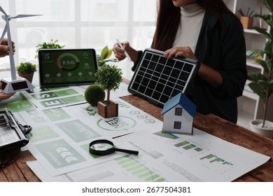 Engineers working with charts and graphs analyzing net zero and ESG project using miniature house, solar panel and wind turbine