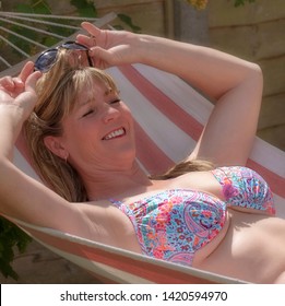 England, UK. June 2017. Woman Wearing A Bikini Top Sunbathing On A Canvas Garden Hammock.