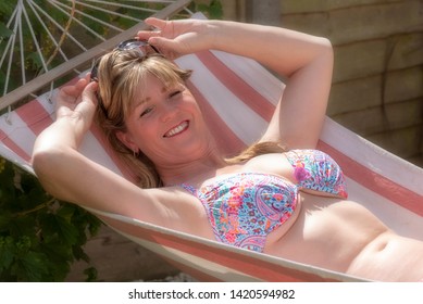 England, UK. June 2017. Woman wearing a bikini top sunbathing on a canvas garden hammock.