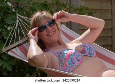 England, UK. June 2017. Woman Wearing A Bikini Top Sunbathing In A Canvas Garden Hammock