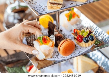 English Afternoon Tea delights with sweet breakfast snacks. Ceremony showcases beautiful desserts and treats. A woman hand selects a dessert from the tea stand.