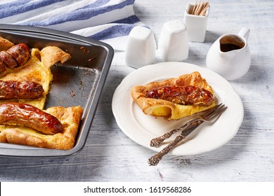 English Classic Dinner: Toad In The Hole Of Baked Yorkshire Pudding With Roasted Sausages Served On A Square Cake Pan And One Portion On A White Plate Served With Onion Gravy, Close-up