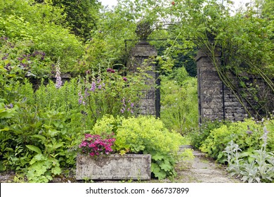 English Cottage Garden With Stone Wall Rose Arch Gate, And Colourful Summer Flowers In Bloom .