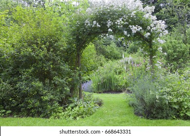 English Cottage Garden With White Rose Arch Entrance, And Colorful Summer Flowers In Bloom .