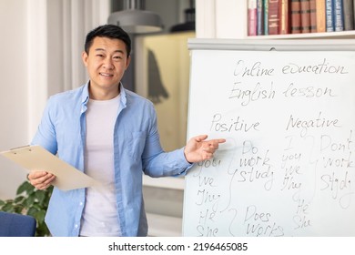 English Course. Happy Asian Male Teacher Having Foreign Language Class Standing Pointing At Whiteboard With Grammar Rules Smiling To Camera Posing In Modern Classroom At School. Education Concept