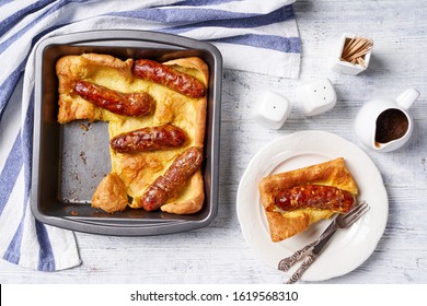 English Cuisine: Toad In The Hole Of Baked Yorkshire Pudding With Roasted Sausages Served On A Square Cake Pan And One Portion On A White Plate Served With Onion Gravy, Horizontal View, Flat Lay