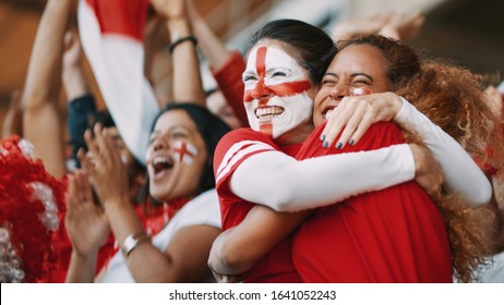 English female soccer fans with England flag painted on their faces hugging each other after their team's victory. English female spectators in football stadium celebrating their team's victory.