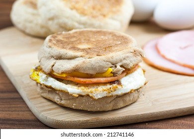 English muffin, egg, ham, and cheese breakfast sandwich on a cutting board with ingredients in the background.