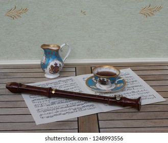 English Teacup With Saucer, Cream Jug, Fine Bone China Porcelain, And A Block Flute On A Sheet Of Music