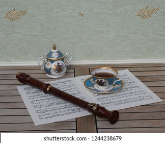 English Teacup With Saucer, Sugar Bowl, Fine Bone China Porcelain, And A Block Flute On A Sheet Of Music